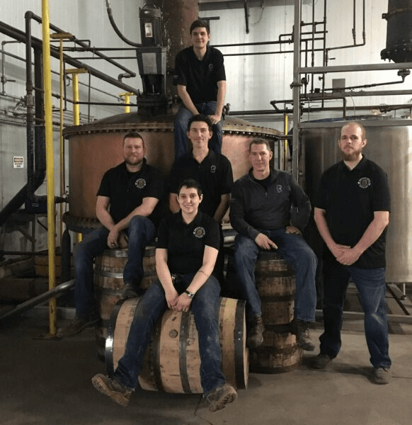 The Kentucky Artisan Distillery team seated on barrels in front of a copper still