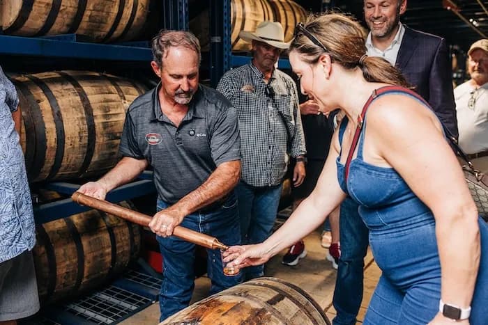 A distillery worker drawing bourbon from a barrel for a visitor